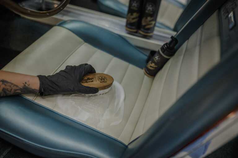 Leather Cleaner – Gros plan main gantée noire avec brosse ronde frottant surface siège cuir crème | Soin Cuir | miio Store