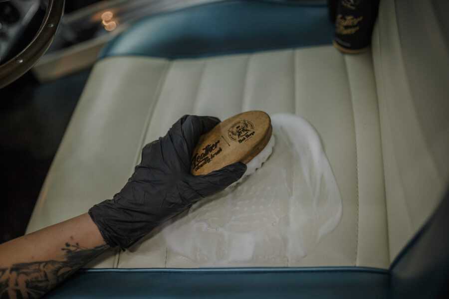 Leather Cleaner – Bras tatoué avec gant noir utilisant brosse ronde sur sièges cuir crème vintage, vue intérieure large | Soin Cuir | miio Store